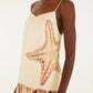 A person wearing the Farm Rio Marine Shells Sand Sleeveless Maxi Dress, featuring a cream hue with a bold starfish motif on the side and a ruffled hem decorated with marine shell patterns, accessorized with gold statement earrings for a striking coastal look.
