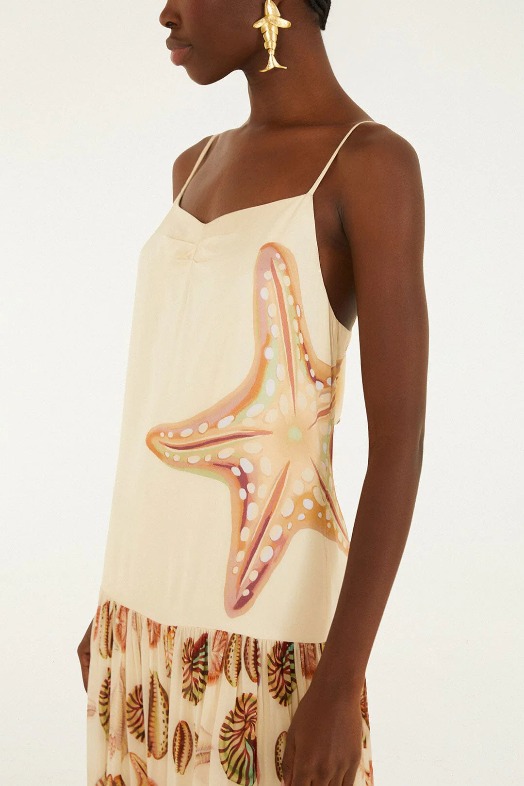A person wearing the Farm Rio Marine Shells Sand Sleeveless Maxi Dress, featuring a cream hue with a bold starfish motif on the side and a ruffled hem decorated with marine shell patterns, accessorized with gold statement earrings for a striking coastal look.