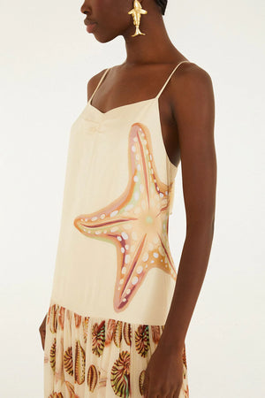 A person wearing the Farm Rio Marine Shells Sand Sleeveless Maxi Dress, featuring a cream hue with a bold starfish motif on the side and a ruffled hem decorated with marine shell patterns, accessorized with gold statement earrings for a striking coastal look.