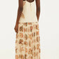 A woman stands facing away, wearing the Farm Rio Marine Shells Sand Sleeveless Maxi Dress—a cream-colored, sleeveless maxi dress with spaghetti straps and a marine shell print on the skirt—paired with flat sandals for a breezy coastal aesthetic.