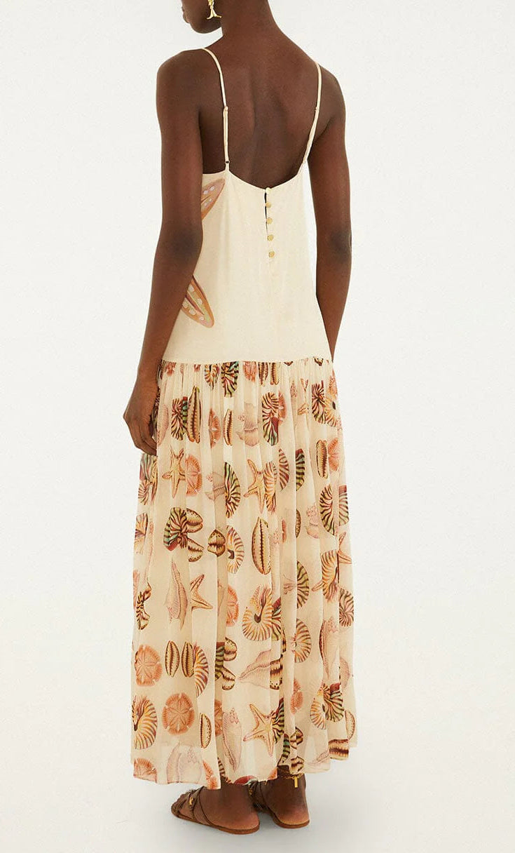 A woman stands facing away, wearing the Farm Rio Marine Shells Sand Sleeveless Maxi Dress—a cream-colored, sleeveless maxi dress with spaghetti straps and a marine shell print on the skirt—paired with flat sandals for a breezy coastal aesthetic.