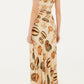 A woman is seen from the back wearing the Farm Rio Marine Shells Sand One Shoulder Maxi Dress, styled with flat sandals for an effortlessly chic look ideal for any summer occasion.