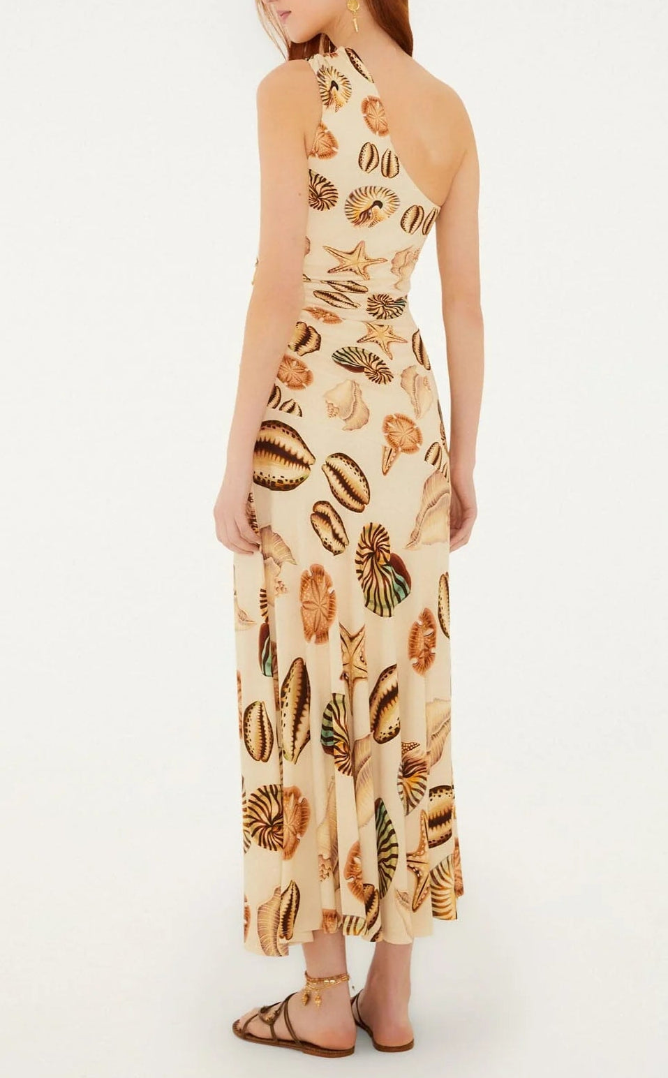 A woman is seen from the back wearing the Farm Rio Marine Shells Sand One Shoulder Maxi Dress, styled with flat sandals for an effortlessly chic look ideal for any summer occasion.