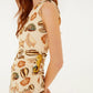 A woman stands sideways wearing the Farm Rio Marine Shells Sand One Shoulder Maxi Dress, featuring a beige hue and marine shell print. Her long, wavy reddish hair and dangling earrings complete this perfect look for any summer occasion.