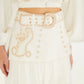 A woman is shown from the torso to upper legs against a plain background, wearing the Farm Rio Sand Richilieu Mini Skirt—featuring beige embroidery, decorative buttons, a large belt—and a matching embroidered top.