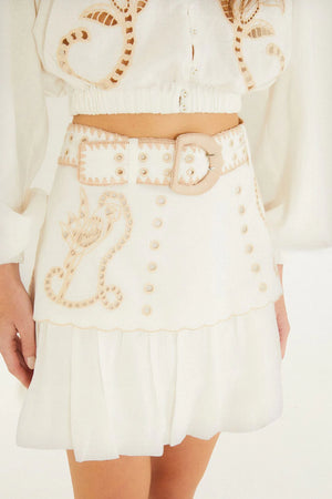 A woman is shown from the torso to upper legs against a plain background, wearing the Farm Rio Sand Richilieu Mini Skirt—featuring beige embroidery, decorative buttons, a large belt—and a matching embroidered top.
