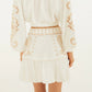 A woman stands facing away, wearing a white long-sleeve blouse paired with the Farm Rio Sand Richilieu Mini Skirt—a belted mini skirt featuring beige embroidery, matching circular accents, and a graceful ruffled hem.