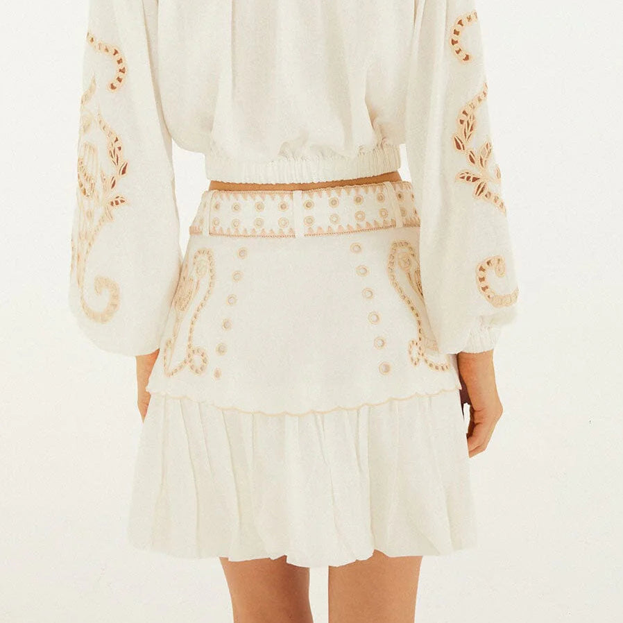 A woman stands facing away, wearing a white long-sleeve blouse paired with the Farm Rio Sand Richilieu Mini Skirt—a belted mini skirt featuring beige embroidery, matching circular accents, and a graceful ruffled hem.
