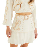 A woman wearing Farm Rio's Sand Richilieu Mini Skirt, a white two-piece set featuring beige embroidery, a belted mini skirt with a scalloped hem, and decorative patterns on both the top and skirt.
