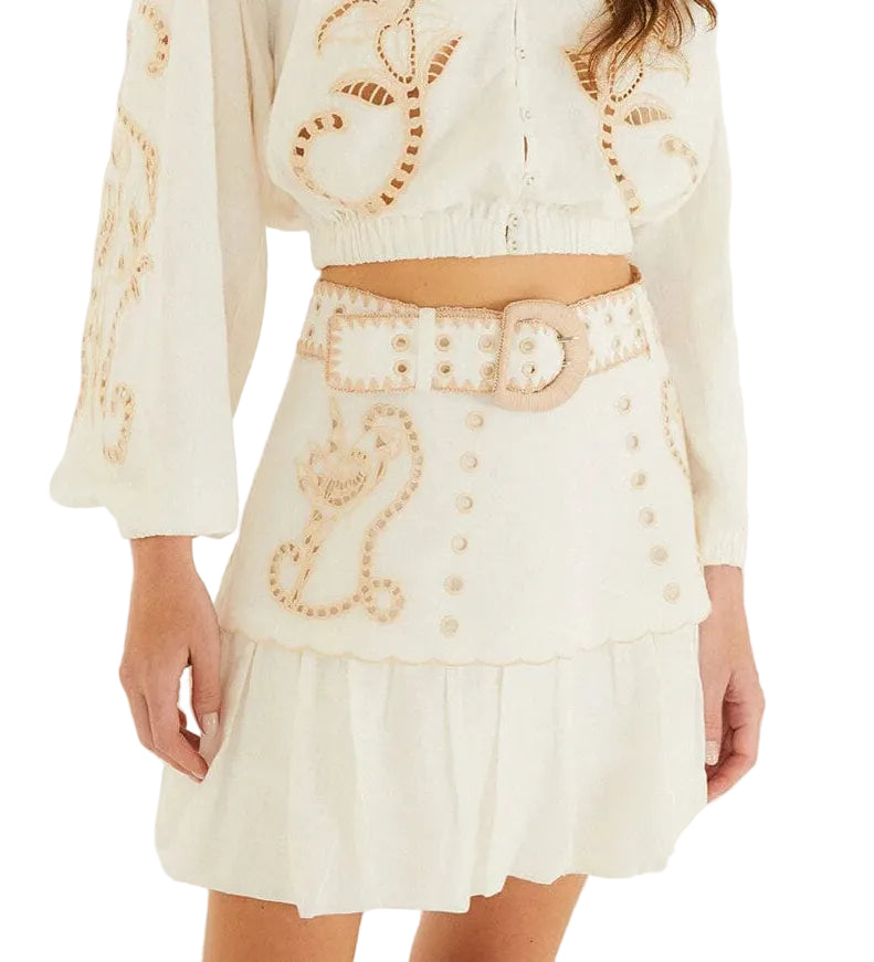 A woman wearing Farm Rio's Sand Richilieu Mini Skirt, a white two-piece set featuring beige embroidery, a belted mini skirt with a scalloped hem, and decorative patterns on both the top and skirt.