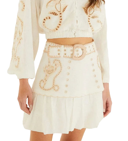 A woman wearing Farm Rio's Sand Richilieu Mini Skirt, a white two-piece set featuring beige embroidery, a belted mini skirt with a scalloped hem, and decorative patterns on both the top and skirt.