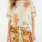 A woman wears the Farm Rio Rio Souvenir Relaxed T Shirt, featuring a cream base with playful seashell and ice cream graphics, styled effortlessly with high-waisted pants adorned with a desert landscape motif for a perfect summer look.