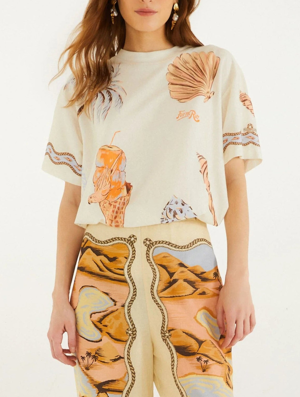 A woman wears the Farm Rio Rio Souvenir Relaxed T Shirt, featuring a cream base with playful seashell and ice cream graphics, styled effortlessly with high-waisted pants adorned with a desert landscape motif for a perfect summer look.