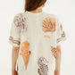 A person with long brown hair is seen from the back wearing the Farm Rio Rio Souvenir Relaxed T Shirt—a cream beach tee featuring playful illustrations of a shell, seahorse, palm leaves, and an ice cream cone with an orange cocktail—making it a perfect pick for effortless summer styling.