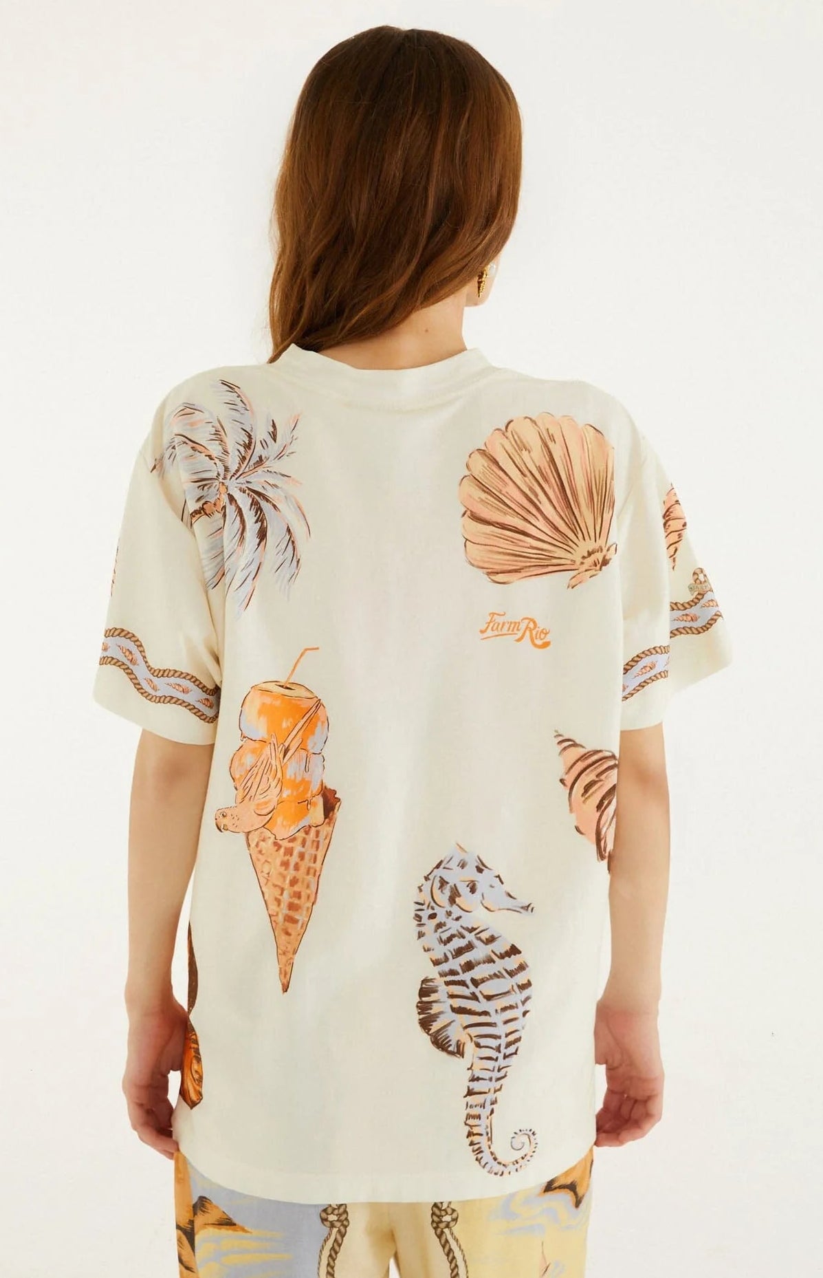A person with long brown hair is seen from the back wearing the Farm Rio Rio Souvenir Relaxed T Shirt—a cream beach tee featuring playful illustrations of a shell, seahorse, palm leaves, and an ice cream cone with an orange cocktail—making it a perfect pick for effortless summer styling.