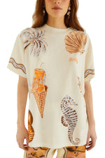 A woman wearing the Farm Rio Rio Souvenir Relaxed T Shirt—a cream beach tee featuring tropical illustrations like a palm tree, seashell, coconut drink, waffle cone, and seahorse—perfect for effortless summer styling. Only her torso and lower face are visible.