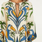 A woman wears the Farm Rio Tropical Graphic Mini Dress featuring a colorful blue, green, yellow, and white botanical pattern, long sleeves, a keyhole neckline with tie, and a playful ruffled hem.