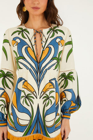 A woman wears the Farm Rio Tropical Graphic Mini Dress featuring a colorful blue, green, yellow, and white botanical pattern, long sleeves, a keyhole neckline with tie, and a playful ruffled hem.