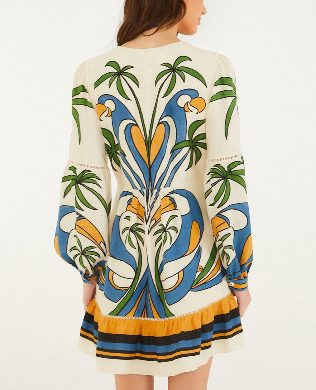 A woman faces away wearing the Farm Rio Tropical Graphic Mini Dress, featuring a cream base with vibrant green, blue, orange, and yellow accents, long sleeves, and a playful ruffled hem.