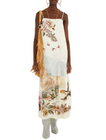 Farm Rio Toile De Rio Off White Maxi Dress featuring a landscape print of buildings, trees, birds, and flowers, styled with a tan fringed shoulder bag and grey boots.