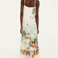 Back view of a person wearing a Farm Rio Toile De Rio Off White Maxi Dress with a tropical landscape and floral print, paired with pale blue heeled boots.