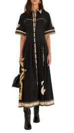 A woman styles the Farm Rio Vintage Souvenirs Black Midi Dress, showcasing its vintage-inspired print, button-down front, and short sleeves. The A-line silhouette creates a flattering look, complemented by black boots and a black handbag.