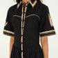 A woman wearing the Farm Rio Vintage Souvenirs Black Midi Dress, which features a vintage-inspired print, collared neckline, gold and white trim, short sleeves, and decorative patterns on the sleeves and skirt. Only her torso and part of her face are visible.