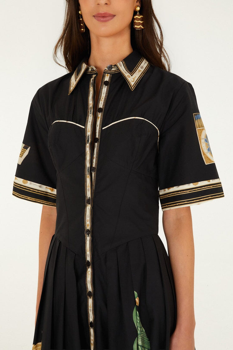 A woman wearing the Farm Rio Vintage Souvenirs Black Midi Dress, which features a vintage-inspired print, collared neckline, gold and white trim, short sleeves, and decorative patterns on the sleeves and skirt. Only her torso and part of her face are visible.
