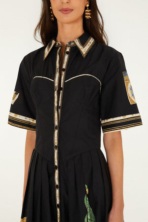 A woman wearing the Farm Rio Vintage Souvenirs Black Midi Dress, which features a vintage-inspired print, collared neckline, gold and white trim, short sleeves, and decorative patterns on the sleeves and skirt. Only her torso and part of her face are visible.