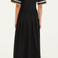 A woman stands facing away, wearing the Farm Rio Vintage Souvenirs Black Midi Dress, which features gold trim and decorative patches on the sleeves, an elegant A-line cut, and is paired with black boots.
