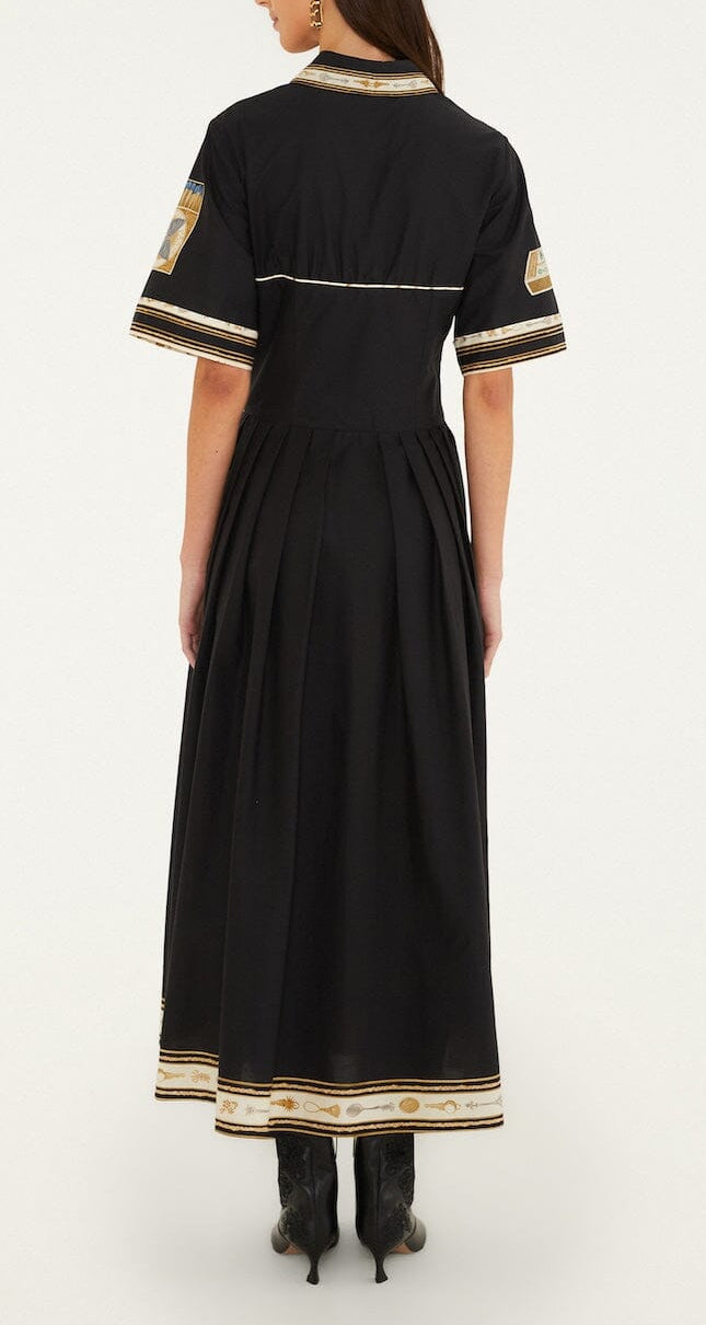 A woman stands facing away, wearing the Farm Rio Vintage Souvenirs Black Midi Dress, which features gold trim and decorative patches on the sleeves, an elegant A-line cut, and is paired with black boots.