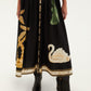 A person stands wearing the Farm Rio Vintage Souvenirs Black Midi Dress, featuring an A-line cut and decorated with gold and green patterns—including a swan—next to black heeled boots on a white background.