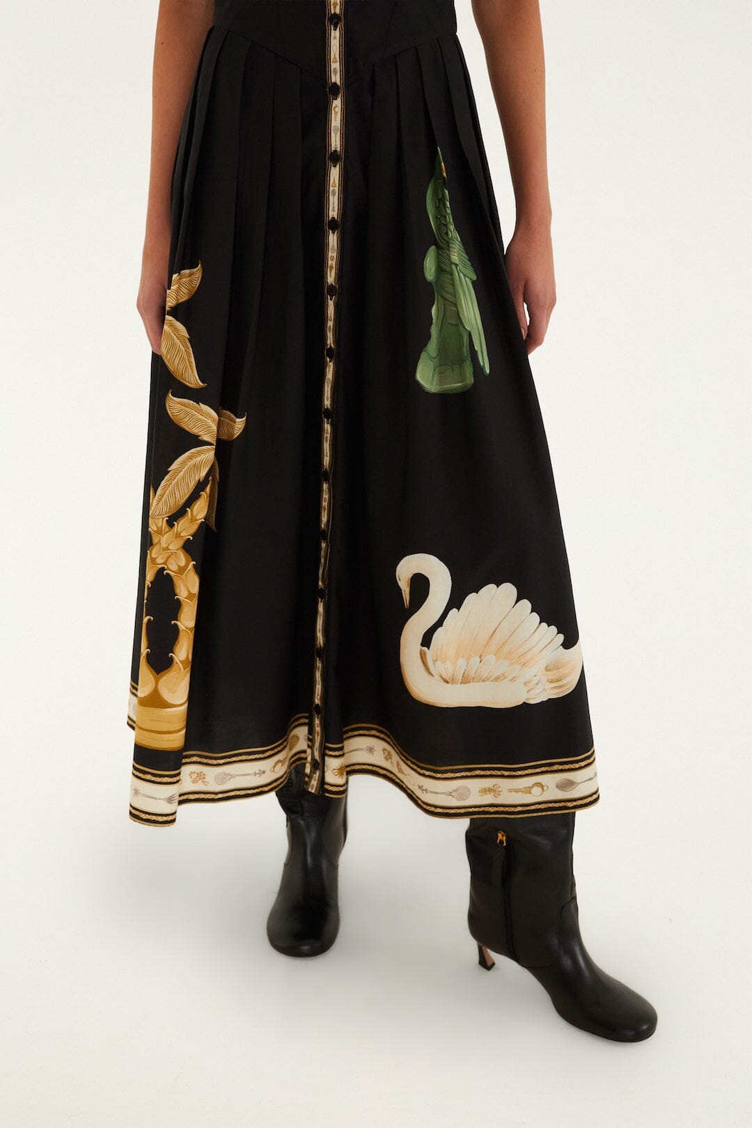 A person stands wearing the Farm Rio Vintage Souvenirs Black Midi Dress, featuring an A-line cut and decorated with gold and green patterns—including a swan—next to black heeled boots on a white background.