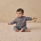 A baby sits on a textured beige rug, wearing the Quincy Mae Baby Boys’ and Boys’ Pocket Sweatshirt in grey pointelle fabric, holding a wooden toy with another toy placed nearby.