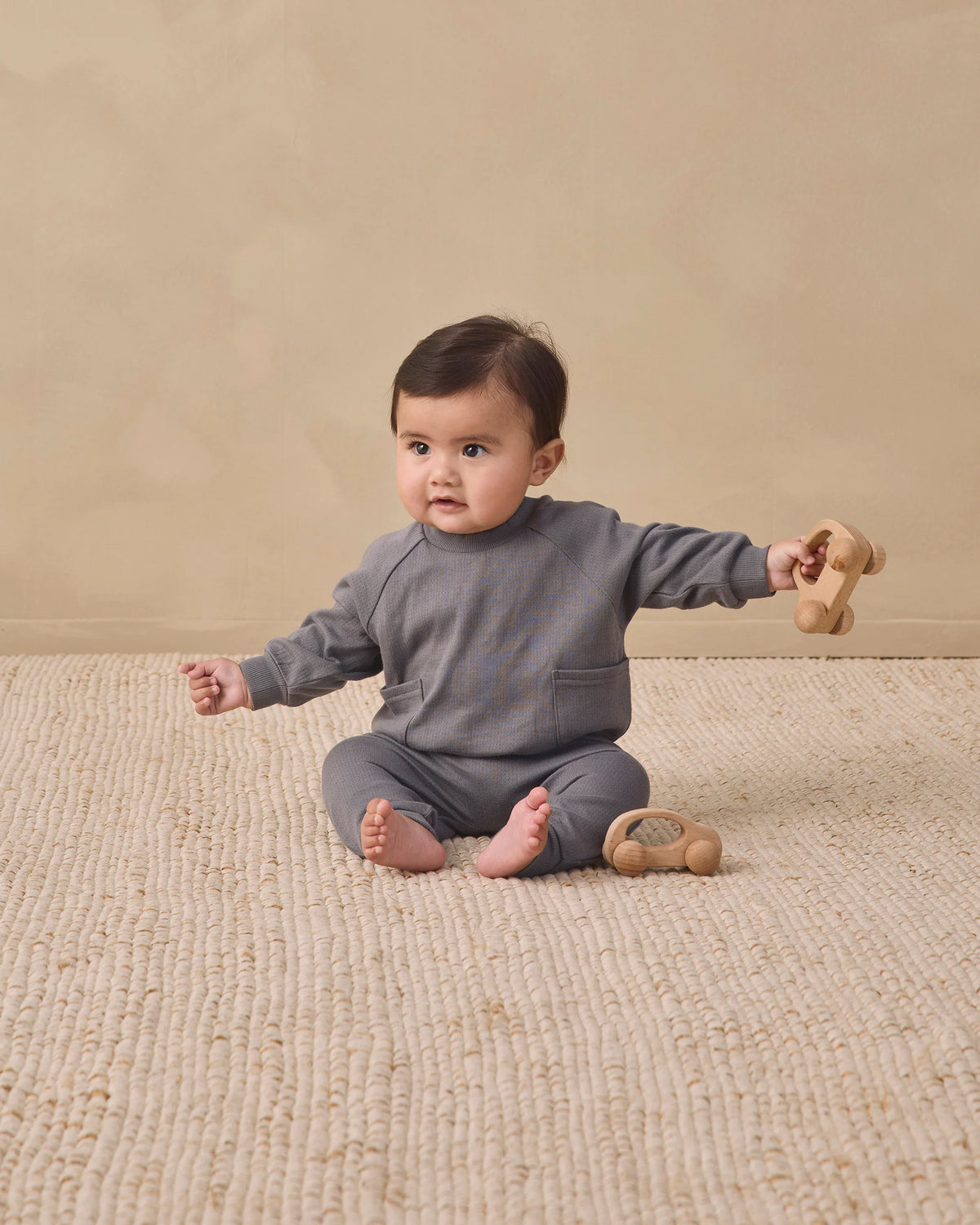 A baby sits on a textured beige rug, wearing the Quincy Mae Baby Boys’ and Boys’ Pocket Sweatshirt in grey pointelle fabric, holding a wooden toy with another toy placed nearby.