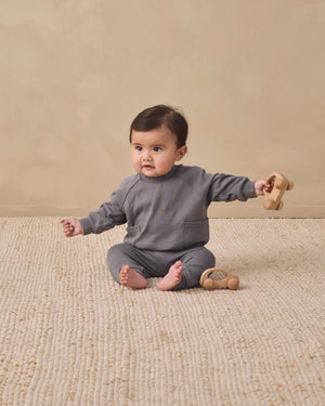 A baby sits on a textured beige rug, wearing the Quincy Mae Baby Boys’ and Boys’ Pocket Sweatshirt in grey pointelle fabric, holding a wooden toy with another toy placed nearby.