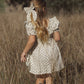 A young girl with long blonde hair adorned with the Girls’ Oversized Bow by Rylee & Cru stands in a field of tall grass, wearing a light floral dress and black boots, facing away from the camera.
