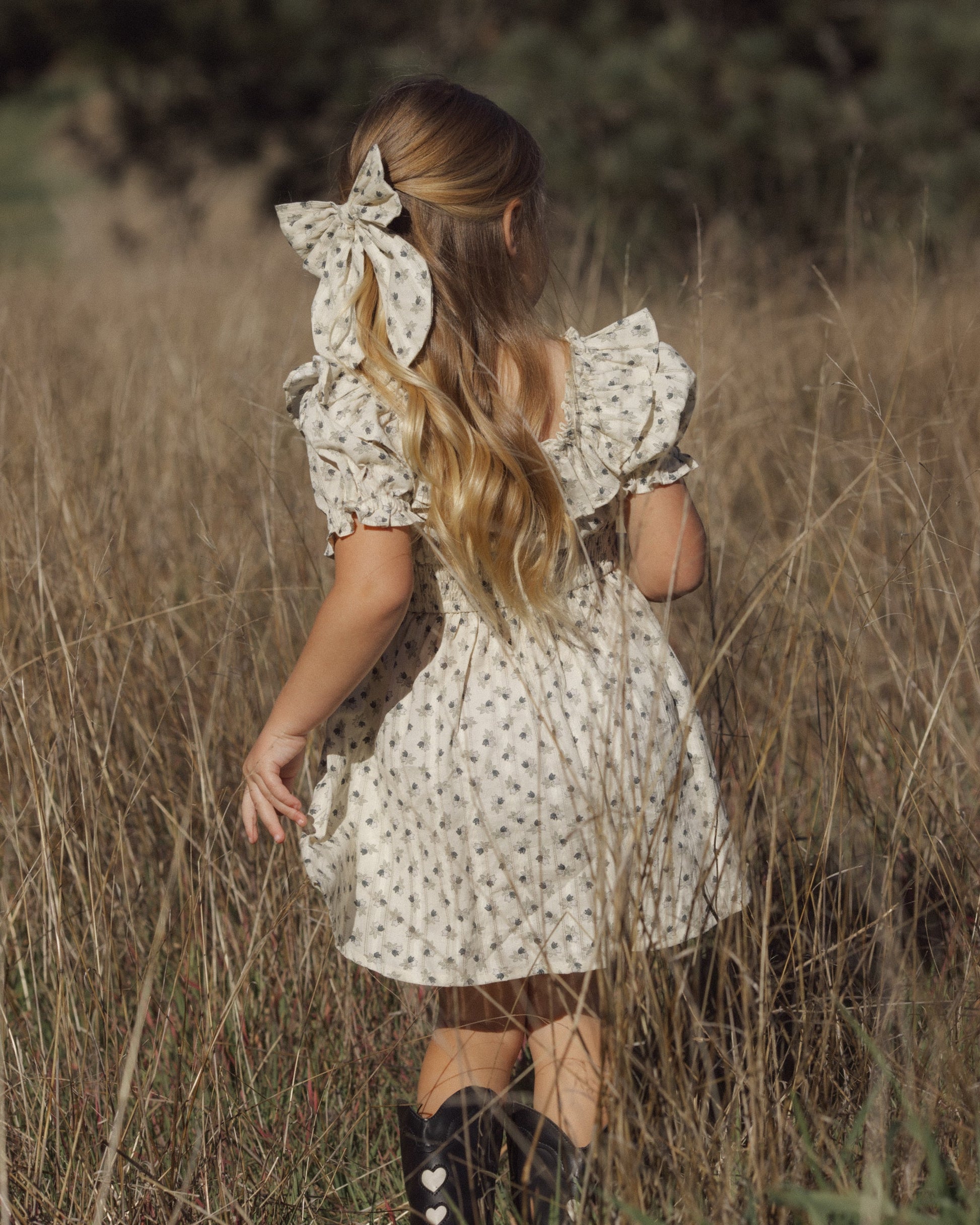 A young girl with long blonde hair adorned with the Girls’ Oversized Bow by Rylee & Cru stands in a field of tall grass, wearing a light floral dress and black boots, facing away from the camera.