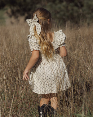 A young girl with long blonde hair adorned with the Girls’ Oversized Bow by Rylee & Cru stands in a field of tall grass, wearing a light floral dress and black boots, facing away from the camera.