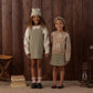 Two young girls stand side by side, holding hands and smiling, each dressed in earth-toned outfits with floral patterns and wrapped in the cozy comfort of the Rylee & Cru Baby Girls’ and Girls’ Aspen Sweater, set against a wooden wall backdrop.