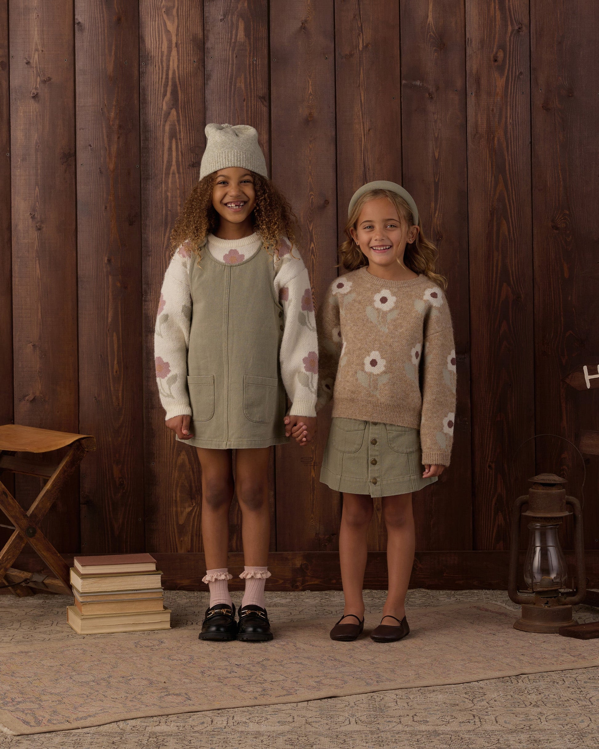 Two young girls stand side by side, holding hands and smiling, each dressed in earth-toned outfits with floral patterns and wrapped in the cozy comfort of the Rylee & Cru Baby Girls’ and Girls’ Aspen Sweater, set against a wooden wall backdrop.