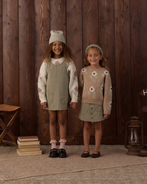 Two young girls stand side by side, holding hands and smiling, each dressed in earth-toned outfits with floral patterns and wrapped in the cozy comfort of the Rylee & Cru Baby Girls’ and Girls’ Aspen Sweater, set against a wooden wall backdrop.