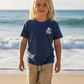 A young child with long blonde hair stands on a sandy beach, wearing the Feather 4 Arrow Baby Boys’ and Boys’ Deep Sea Adventures Vintage Tee in navy paired with tan shorts, with waves and the ocean in the background.