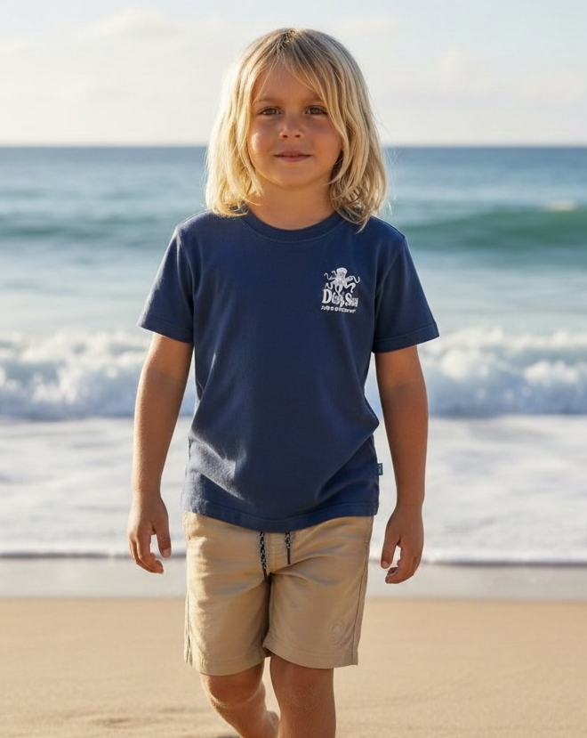 A young child with long blonde hair stands on a sandy beach, wearing the Feather 4 Arrow Baby Boys’ and Boys’ Deep Sea Adventures Vintage Tee in navy paired with tan shorts, with waves and the ocean in the background.