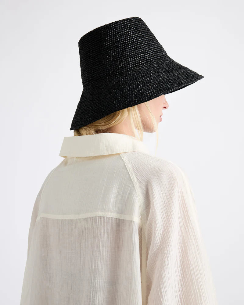 A person wearing a sheer, cream-colored shirt and the Felix Hat by Janessa Leone, a wide-brimmed black summer hat, shown from the back and side.