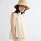 A woman stands against a plain white background wearing a sleeveless, light beige halter top and the Janessa Leone Felix Hat, a chic raffia bucket hat.