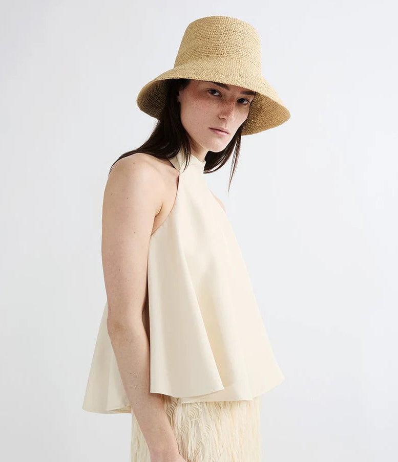 A woman stands against a plain white background wearing a sleeveless, light beige halter top and the Janessa Leone Felix Hat, a chic raffia bucket hat.