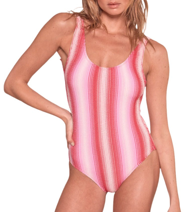 A person poses confidently in the Lemlem Haset One Piece, a pink and red striped swimsuit crafted from scrunch rib fabric. With one hand on their hip, they exude vibrant charm against a plain backdrop, embodying the signature style of Lemlem swimwear.