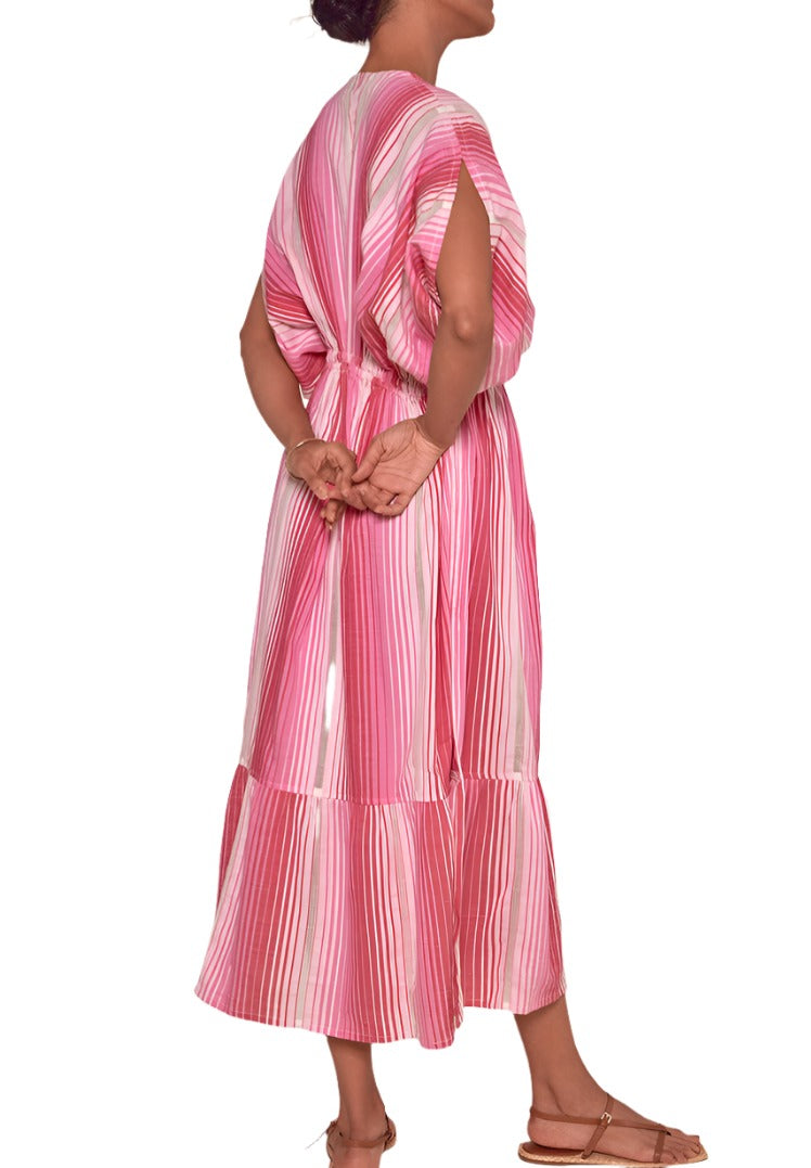 A person in a Lemlem Leila Plunge Dress, hand-woven in Ethiopia from 100% cotton with pink and white stripes, stands with their back turned and hands clasped behind their back.