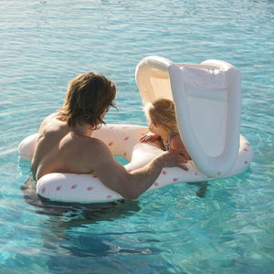 An adult and a young child are in a pool with clear, blue water. The adult holds the Sunnylife Strawberry Sunshine Float Together Baby Seat, which features a removable shade canopy, while the child sits comfortably inside it.